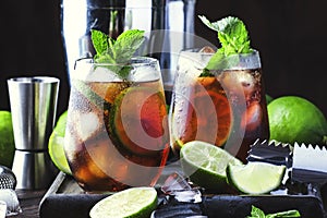 Cuba libre alcohol cocktail with golden rum, lemon juice, cola, lime and ice, dark bar counter background, bar tools, place for