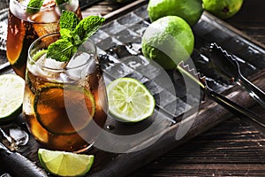Cuba libre alcohol cocktail with golden rum, lemon juice, cola, lime and ice, dark bar counter background, bar tools, place for