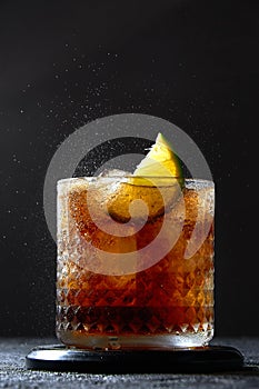 Cuba Libre or Long Island Cocktail with rum, cola and lime in glass. Soda spray on dark background. Refreshing drink