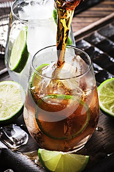 Cuba libre preparation alcohol cocktail with golden rum, lemon juice, cola, lime and ice, dark bar counter background, bar tools,