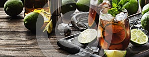 Cuba libre or rum cola cocktail with strong alcohol, lemon juice, lime and ice, dark background, steel bar tools, copy space