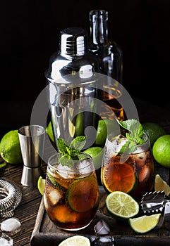 Cuba libre or rum cola cocktail with strong alcohol, lemon juice, lime and ice, dark background, steel bar tools, copy space