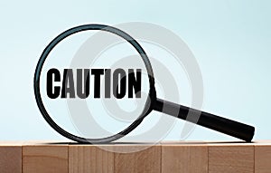 Cubes on a light blue wooden background. On them a magnifying glass with the word CAUTION
