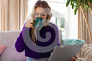 Cucasian woman using laptop and drinking coffee, sitting on couch working at home