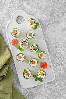 Cucumbers rolls with cream cheese, salted salmon and fresh herbs served on a white ceramic board on a concrete background.