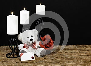 Cudlely teddy bear with red bow tie, red rose, white candles perched on black candle holders on mesh place mat and wooden table wi