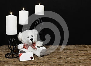 Cudlely teddy bear with red bow tie, white candles perched on black candle holders on mesh place mat and wooden table with card an