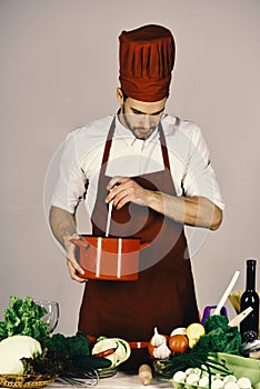 Cuisine and professional cooking concept. Chef with busy face holds red saucepan on grey background.