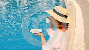 The culture of drinking tea and coffee. Woman with straw hat drinking hot drink from a cup near blue swimming pool