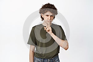 Cunning young man shushing, making hush sign on lips and smiling, make shh gesture, standing over white background