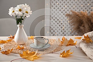 Cup of aromatic coffee, beautiful flowers and autumn leaves on white wooden table indoors, space for text