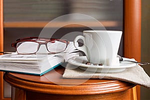 A cup with aromatic coffee, a volume of poetry, women`s reading glasses on a wooden console at home.