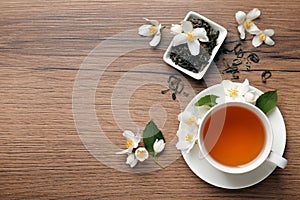 Cup of aromatic jasmine tea, dry leaves and fresh flowers on wooden table, flat lay. Space for text