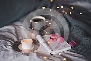 Cup of blac tea with scented burning candle with open paper book and pink fruit macaroons in bed on knit jumper over glow lights a