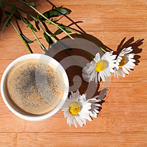 A cup of black coffee branch of white daisy flowers on wooden background in morning bright sunligh with shadow