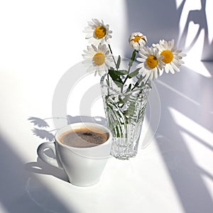 A cup of black coffee with foam, a bouquet of white camomile flowers in a crystal vase with water on a white table in the sunlight