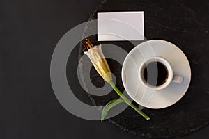 Cup of black coffee and fresh spring bud of lily flower with clean white business card wihhout massage on a round slate on black