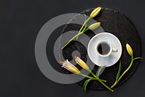 Cup of black coffee and fresh spring buds of lily flowers on a round slate on a black background.Romantic stylish dating breakfast