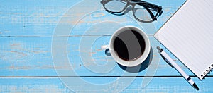 Cup of black coffee with office supplies; pen, notebook and eyes glasses on blue wooden table background