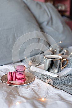 Cup of black coffee with paper book and burning candle with pink tasty macarons cookies in bed close up.