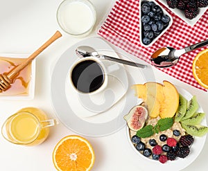 Cup of black coffee, a plate of oatmeal and fruit, honey and a glass of milk on a white table, healthy morning breakfast, top view