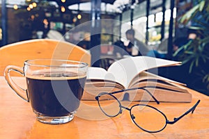 A cup of black coffee with reading glasses on table and open boo