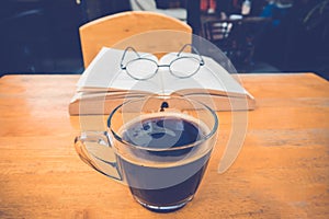 A cup of black coffee with reading glasses on table and open boo