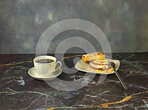 A cup of black coffee on a saucer and a plate with two cinnamon rolls and a fork are on the marble table