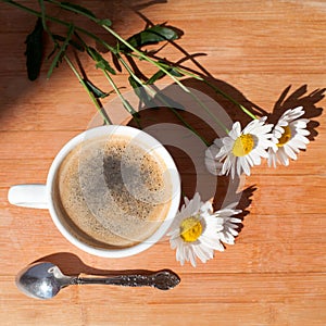 A cup of black coffee, silver spoon, branch of white daisy flowers on wooden background top view