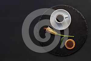 Cup of black coffee tea, cookie with chocolate, single bud of lily flower on a round slate on a black background