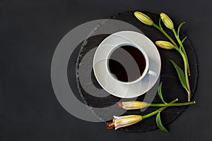 Cup of black coffee tea and fresh spring buds of lily flowers on a round slate on a black background. Romantic stylish breakfast.