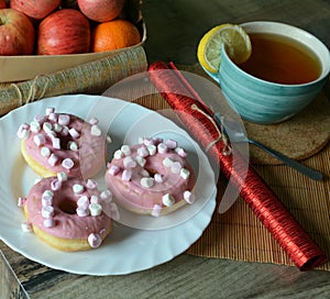 Cup of black tea with lemon and three donuts with pink icing on a plate on a wooden table. Sweet breakfast.