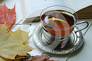 A Cup of black tea with lemon on the windowsill on the background of autumn leaves and an open book close-up