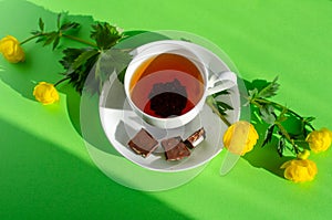 A Cup of black tea, pieces of chocolate and yellow flowers of Trollius europaeus on a green background. Chocolate day