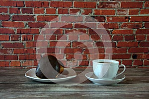A cup of black tea on a saucer and a plate with two donuts in chocolate icing lie on a wooden table against a brick wall. Close-up