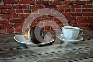 A cup of black tea on a saucer and a plate with two donuts in chocolate icing lie on a wooden table against a brick wall. Close-up