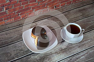 A cup of black tea on a saucer and a plate with two donuts in chocolate icing lie on a wooden table against a brick wall. Close-up