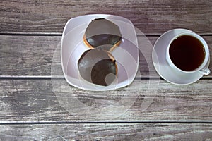 A cup of black tea on a saucer and a plate with two donuts in chocolate icing lie on a wooden table. Close-up