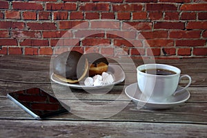 A cup of black tea on a saucer and a plate with two donuts in chocolate icing and pieces of bize, are lying on a wooden table