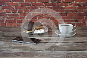 A cup of black tea on a saucer and a plate with two donuts in chocolate icing and pieces of bize, are lying on a wooden table