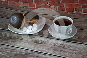 A cup of black tea on a saucer and a plate with two donuts in chocolate icing and pieces of bize, are lying on a wooden table