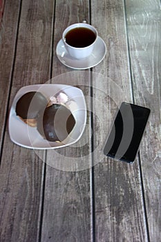 A cup of black tea on a saucer and a plate with two donuts in chocolate icing and pieces of bize, are lying on a wooden table wall
