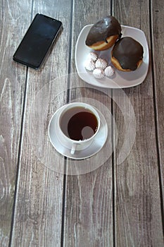 A cup of black tea on a saucer and a plate with two donuts in chocolate icing and pieces of bize, are lying on a wooden table wall