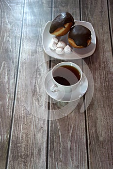 A cup of black tea on a saucer and a plate with two donuts in chocolate icing and pieces of bizet, lie on a wooden table. Close-up