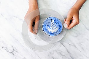 Cup of blue matcha latte in woman hands on white marble table trendy background