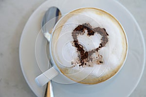 Cup of cappuccino with cocoa powder in heart shape on a table in a street cafe, high angle view from above, selected focus, narrow