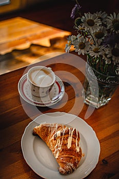 Cup of cappuccino coffee and croissant / brioche on a wooden table with freshly gathered daisy flowers. Italian breakfast.