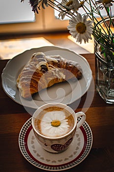 Cup of cappuccino coffee and croissant / brioche on a wooden table with freshly gathered daisy flowers. Italian breakfast.