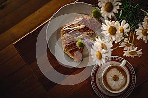 Cup of cappuccino coffee and croissant / brioche on a wooden table with freshly gathered daisy flowers. Italian breakfast.