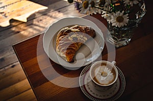 Cup of cappuccino coffee and croissant / brioche on a wooden table with freshly gathered daisy flowers. Italian breakfast.